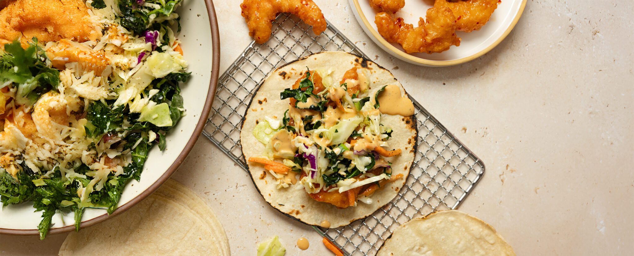 Overhead view of Social Kitchens Sweet & Spicy Shrimp tacos with slaw, served on a wire rack with a bowl of slaw and shrimp pieces off to the side of the tabletop.