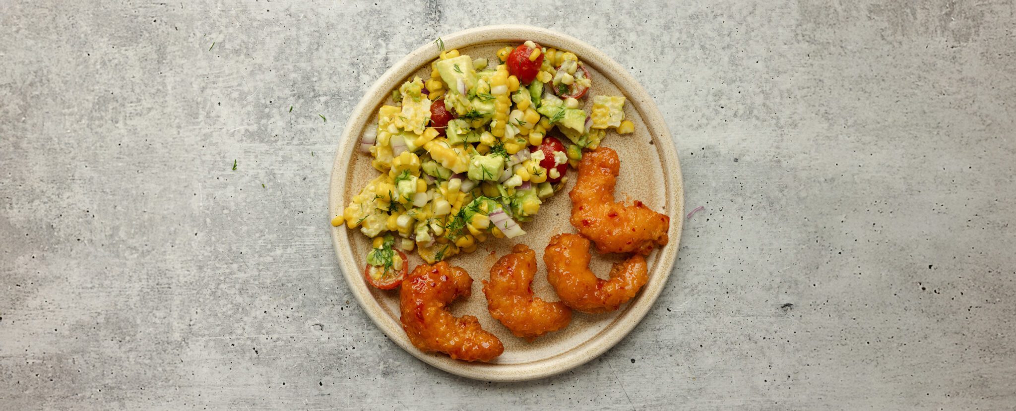 Overhead view of a white ceramic plate of Social Kitchens Sweet & Spicy Shrimp with corn & avocado salsa, served over a textured light gray stone tabletop.