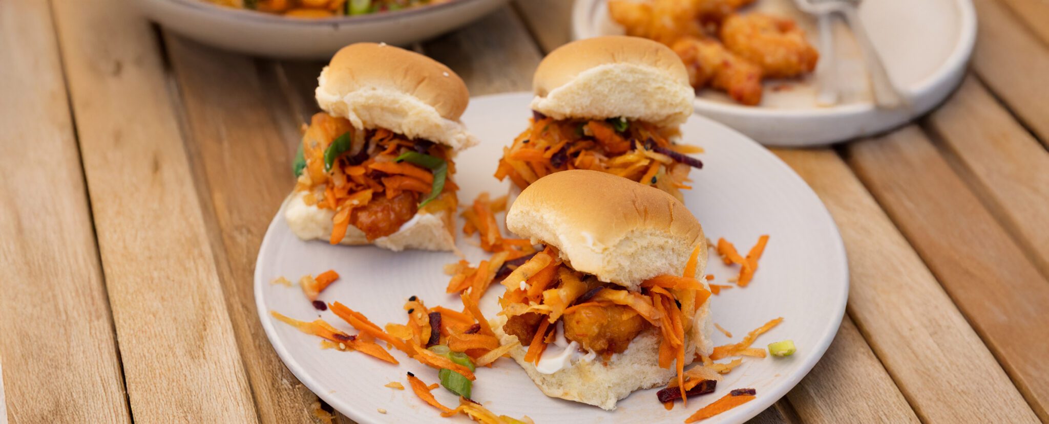 Three crispy shrimp sliders with sesame rainbow salad served on on a white plate over a wooden picnic table with a couple other dishes in the background.
