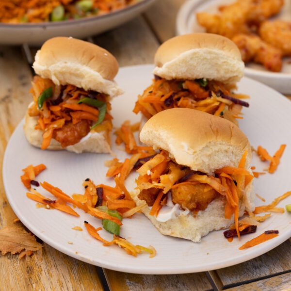 Three crispy shrimp sliders with sesame rainbow salad served on on a white plate over a wooden picnic table with a couple other dishes in the background.