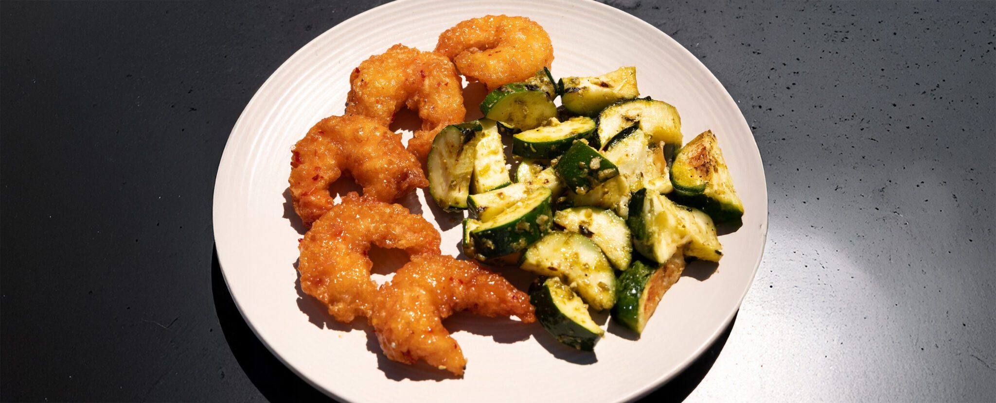 Three-quarter view of a white plate filled with Social Kitchens Sweet & Spicy shrimp and grilled pesto zucchini, served over a black tabletop.