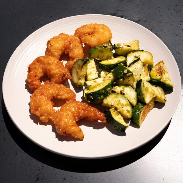 Three-quarter view of a white plate filled with Social Kitchens Sweet & Spicy shrimp and grilled pesto zucchini, served over a black tabletop.