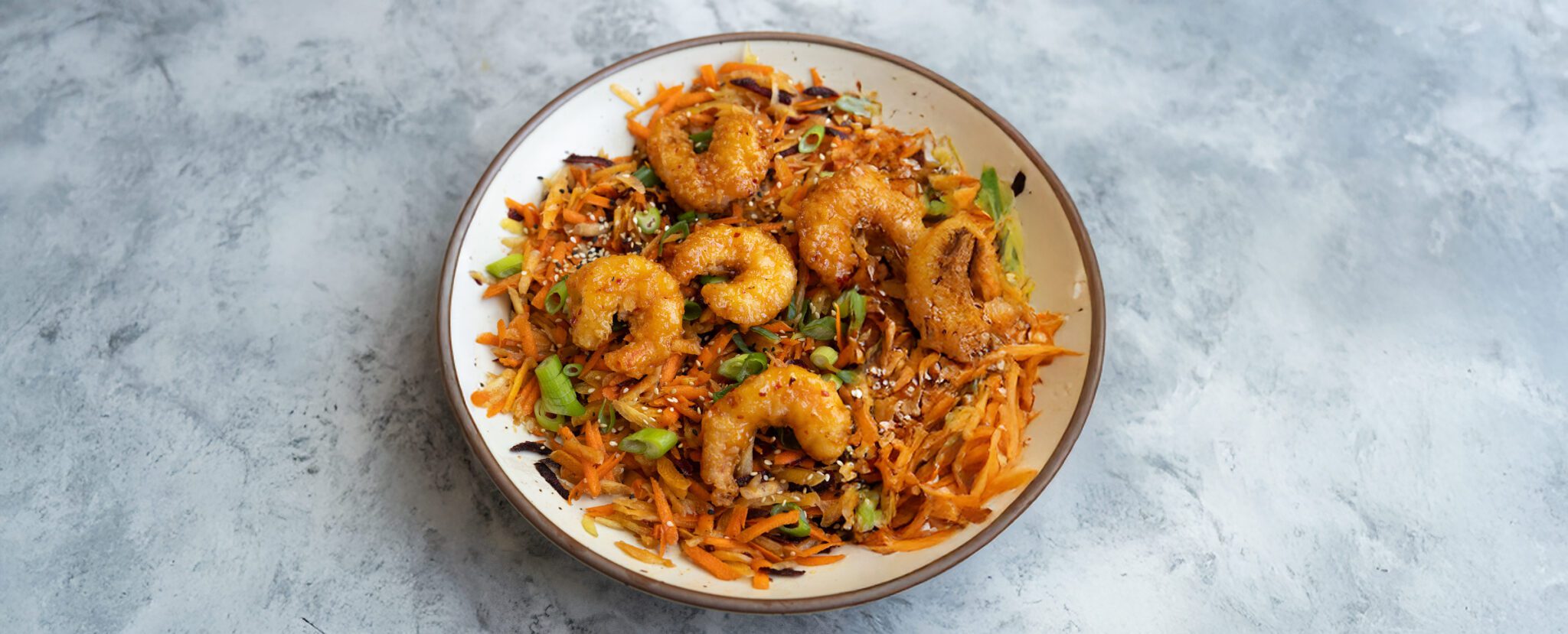 Three-quarter view of a a bowl of sesame rainbow carrot salad with shrimp and garnished with green onions, served over marble tabletop.