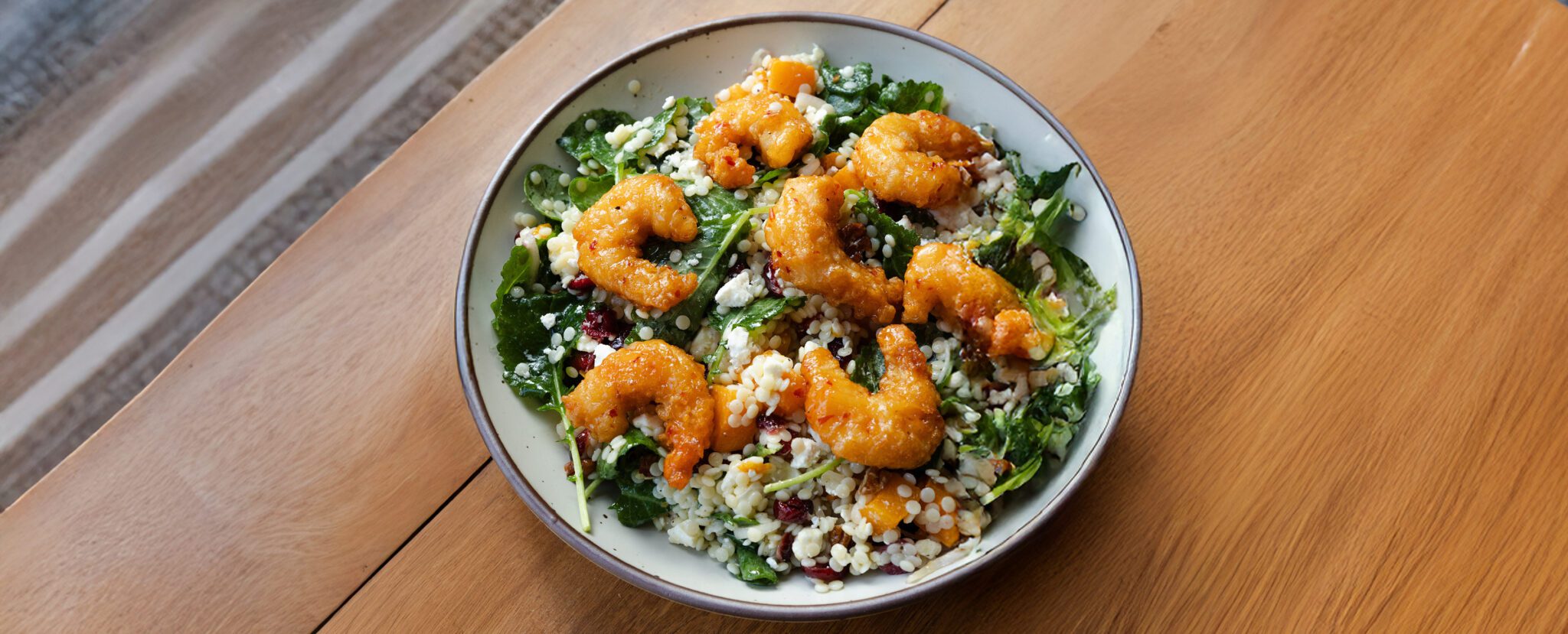 A white plate of fall couscous salad topped with Social Kitchens Sweet & Spicy Shrimp served on a wooden tabletop.