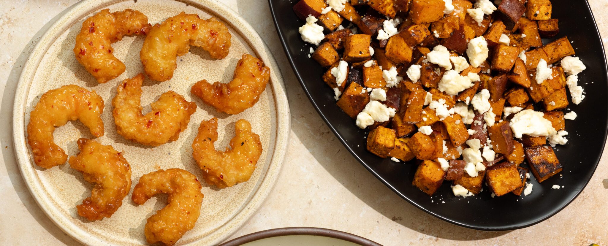 Overhead view of roasted chipotle sweet potatoes with feta on a large black plate and a small white plate of Social Kitchens Sweet & Spicy Shrimp.