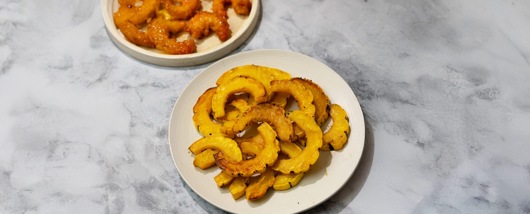 Three-quarter view of roasted maple delicata squash and a separate plate off to the side of Social Kitchens Sweet & Spicy Shrimp, served on a textured white and gray tabletop.
