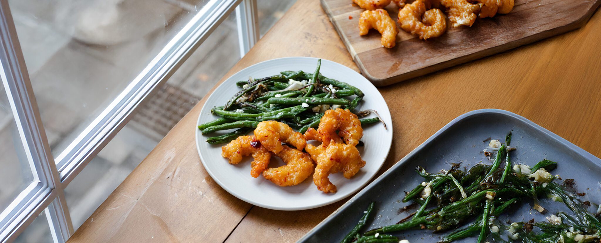 A white plate served with Social Kitchen's Sweet & Spicy Shrimp with green beans placed beside a white-paned window on a wooden tabletop. Off to the side is a small wooden cutting board plated with more shrimp and a blue tray of roasted green beans.