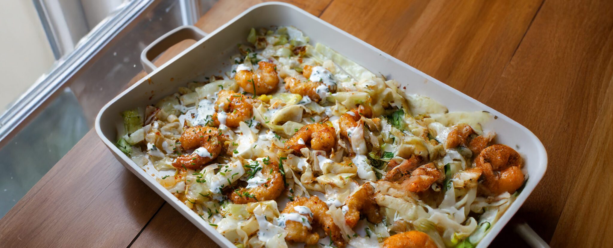 Three-quarter view of Social Kitchens Sweet & Spicy Shrimp with cabbage baked in a deep baking dish with handles, placed on a wooden tabletop near a window.
