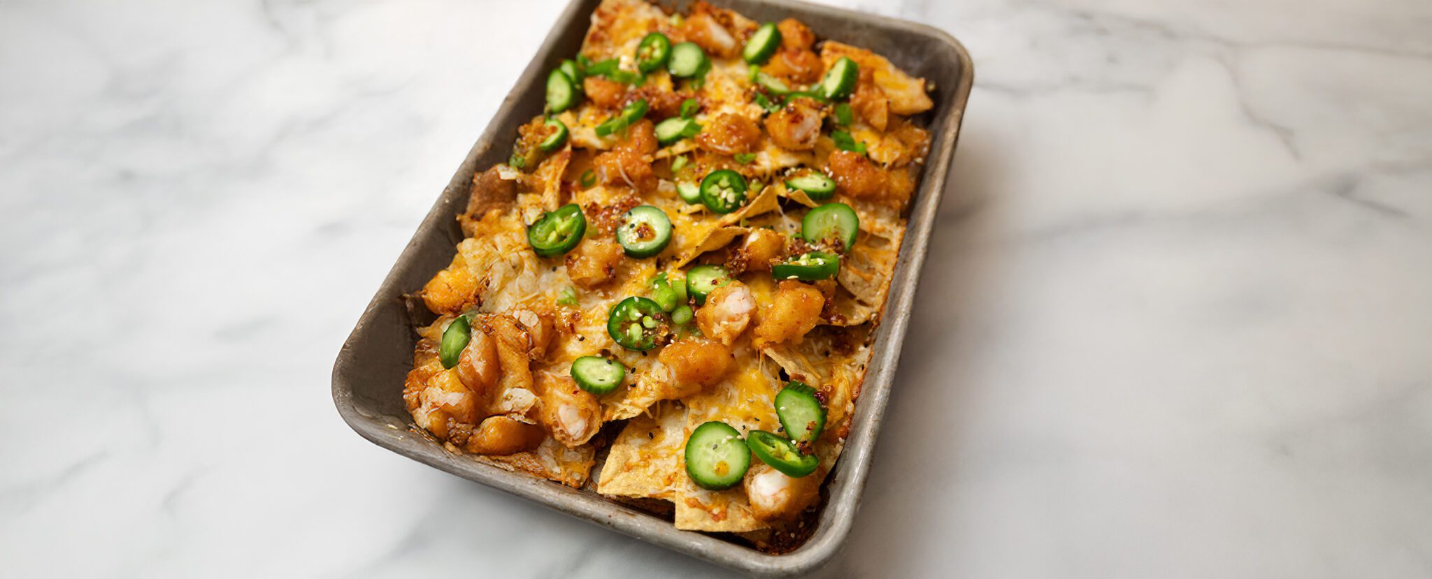 Three-quarter view of a metal sheet pan containing Sweet & Spicy Shrimp nachos garnished on top with jalapeño pepper slices.