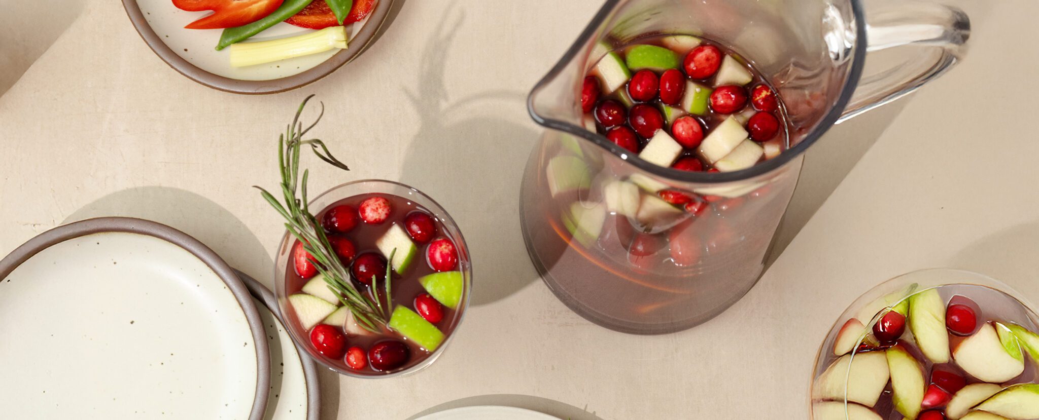 Overhead view of a glass pitcher and several glasses of holiday sangria with green apple pieces, cranberries, some lemon & orange slices, and sprigs of rosemary for garnish.