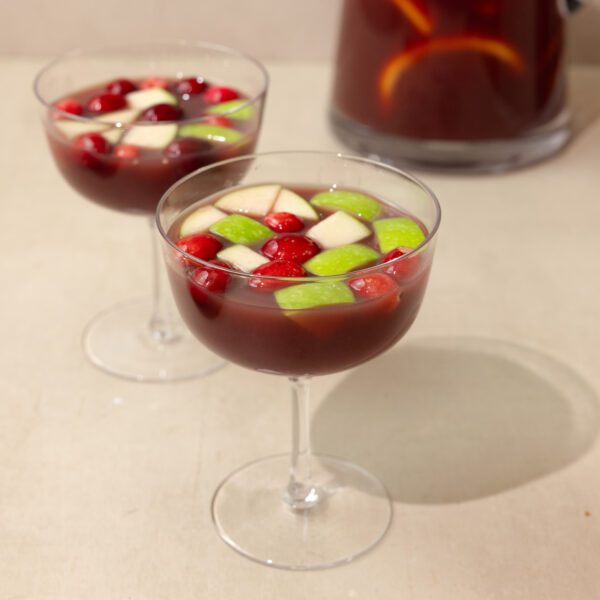 Two wine glasses filled with holiday sangria with green apple pieces and cranberries. A filled pitcher of sangria sits in the background.