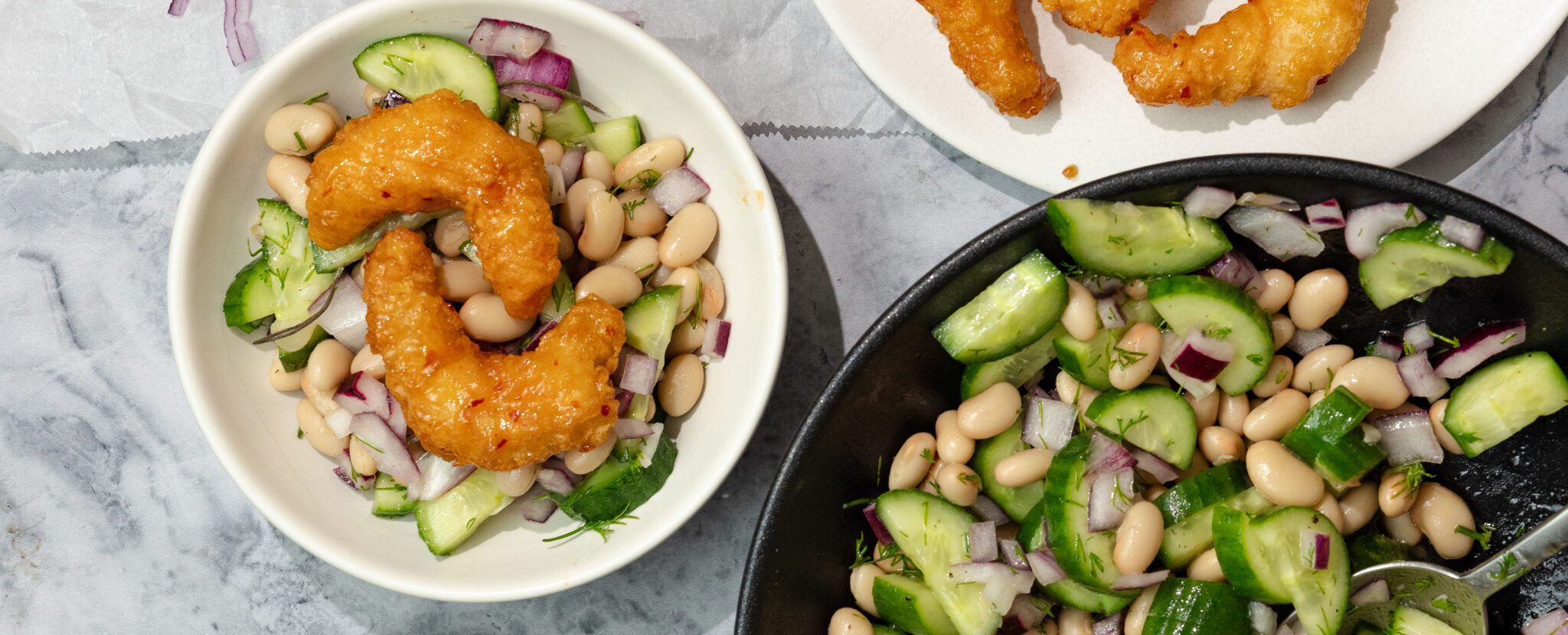 Overhead view of a white plate of Social Kitchens Sweet & Spicy Shrimp with Lemon Dill Bean Salad. Off to the side are more pieces of shrimp and a black bowl of more Lemon Dill Bean Salad.