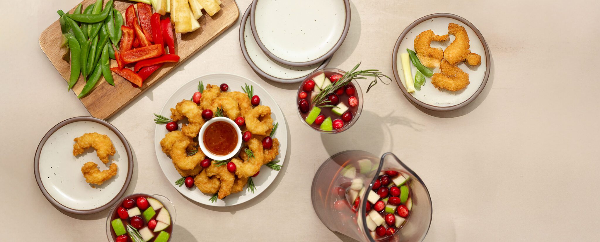 Overhead view of a winter seasonal tablescape of Social Kitchens Sweet & Spicy Shrimp garnished with cranberries and small sprigs of rosemary, a glass and pitcher full of Holiday Sangria, a vegetable charcuterie board, and additional ceramic plates for sharing.