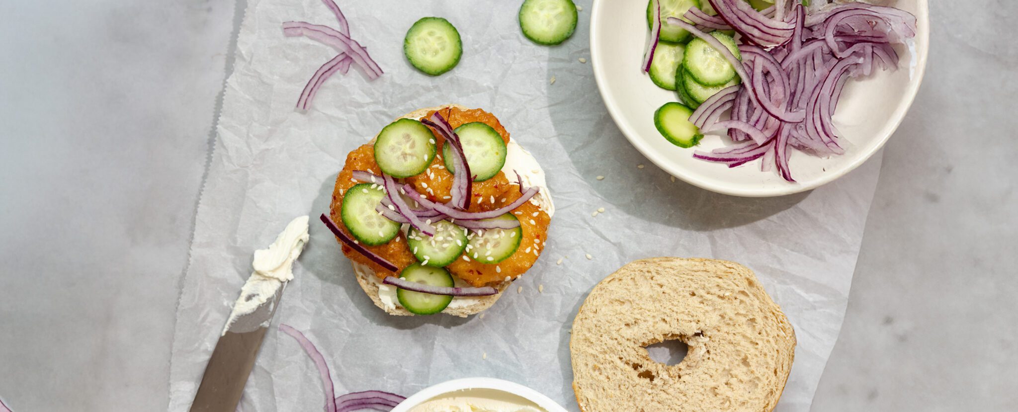 Overhead view of Social Kitchens Sweet & Spicy Shrimp Bagels, garnished with cucumbers and red onion.