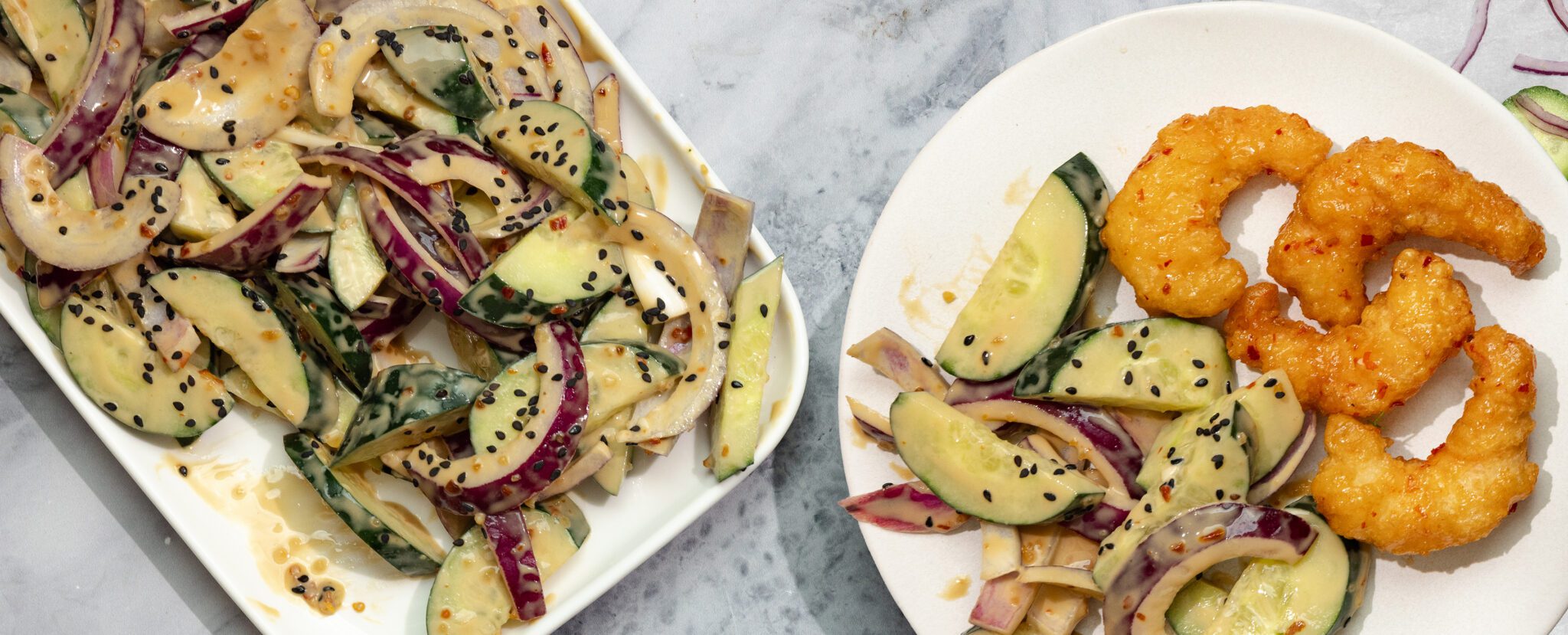 Overhead view of a rectangular plate of Tahini Cucumber Salad and another white plate of salad and Social Kitchens Sweet & Spicy Shrimp, served on a marble tabletop.
