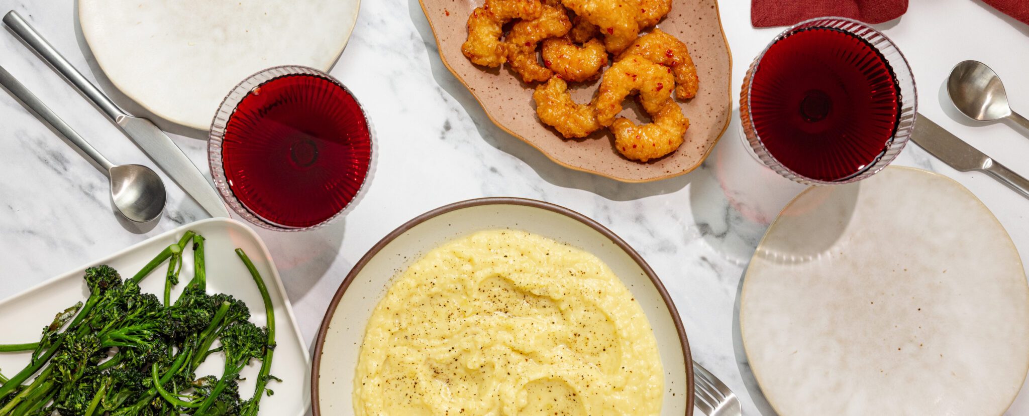 Overhead view of a tablescape containing miso butter mashed potatoes, broccolini, Social Kitchens Sweet & Spicy Shrimp, glasses of wine and a set of tableware all over a marble table surface.