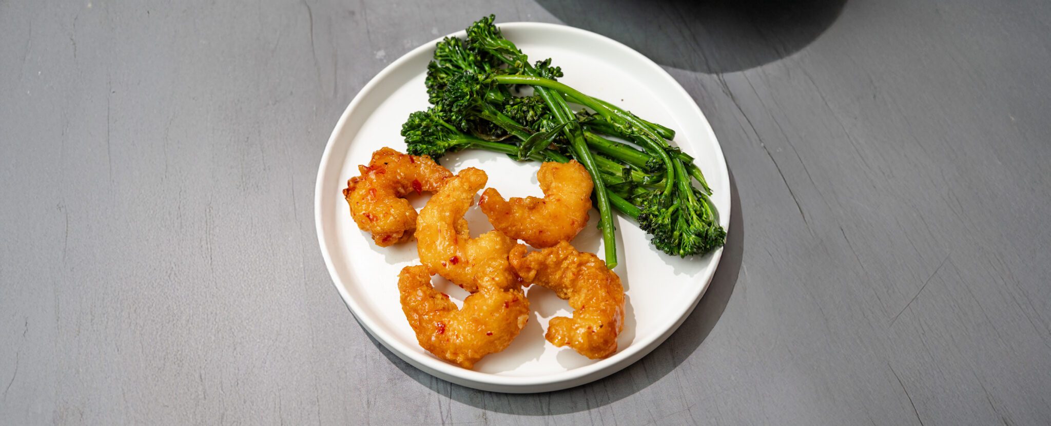 Three-quarter view of Social Kitchens Sweet & Spicy Shrimp with sautéed broccolini on a round plate over a wooden gray tabletop.