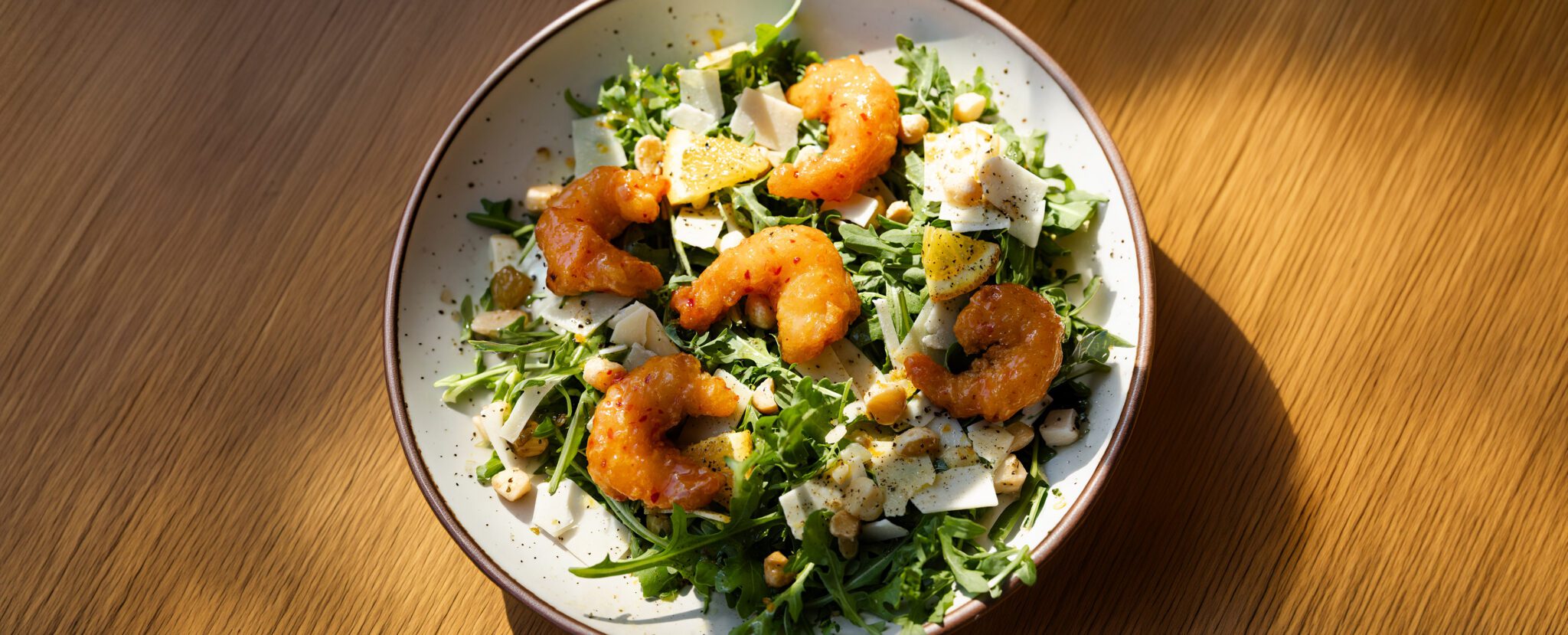 Three-quarter view of a Citrus Arugula Salad in a brown-rimmed ceramic plate, garnished with Social Kitchens shrimp, shaved parmesan, lemon slices, and chopped macadamia nuts.
