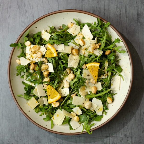 Overhead view of a Citrus Arugula Salad in a brown-rimmed ceramic plate, garnished with shaved parmesan, lemon slices, and chopped macadamia nuts.
