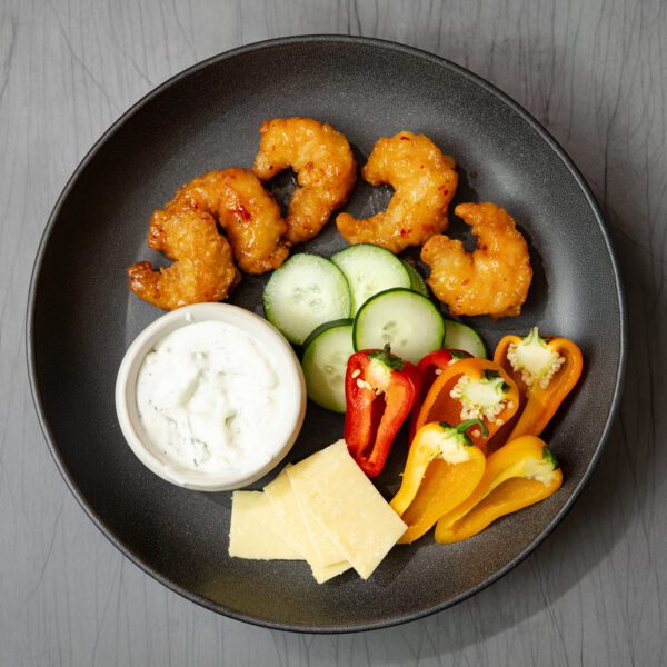 Overhead view of a round black plate arranged with Social Kitchens shrimp, sliced cucumbers, red & yellow bell pepper halves, tzatziki dip, and sliced cheese.