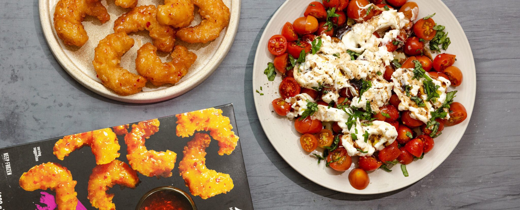 Overhead view of Social Kitchens Sweet & Spicy Shrimp on a white plate and another plate of Burrata Caprese. Off to the side is a box of Social Kitchens shrimp packaging.