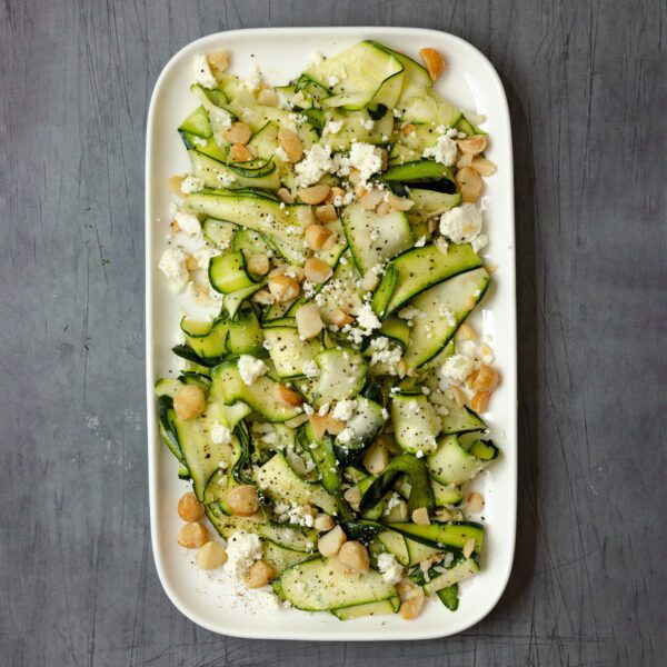 Overhead view of a Shaved Zucchini Salad garnished with crumbled feta and nuts, on a white large rectangular platter over a gray wood tabletop.