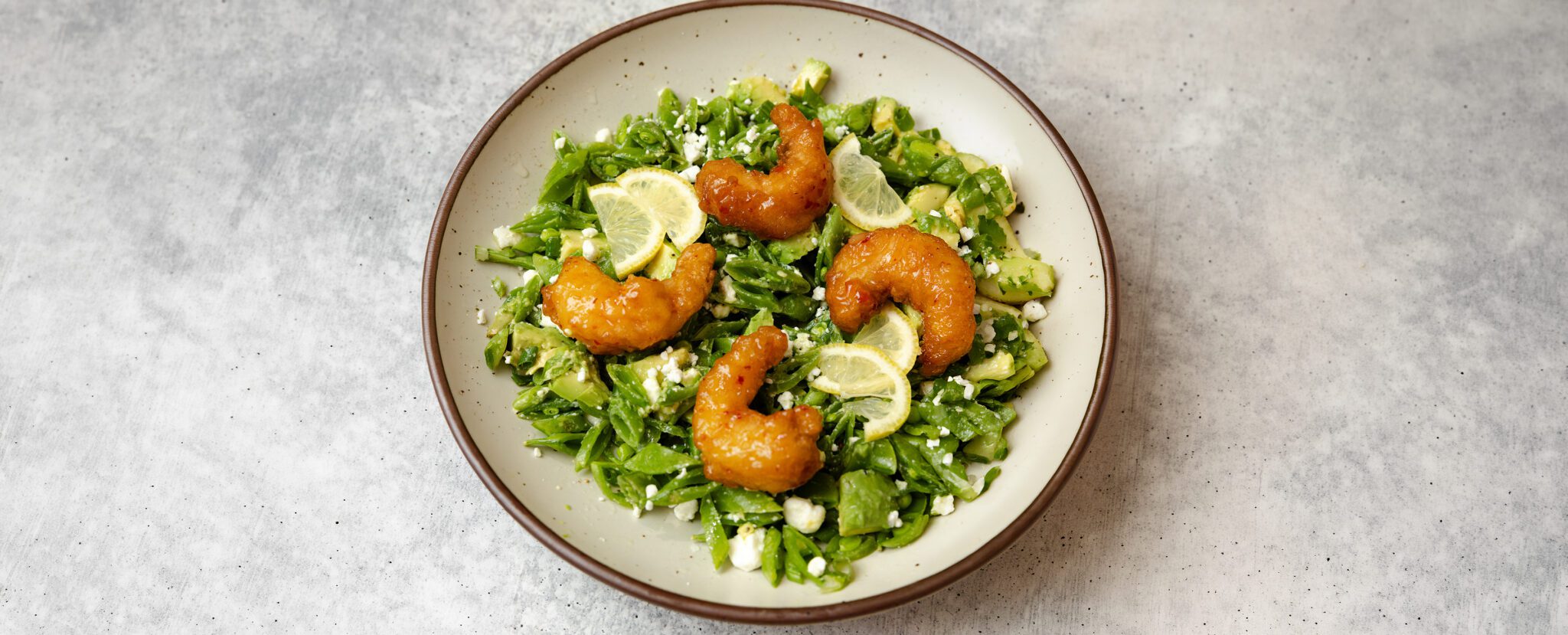 Three-quarter angle view of a Snap Pea & Avocado Salad with Social Kitchens Sweet & Spicy Shrimp on a round plate, served over a textured stone tabletop.