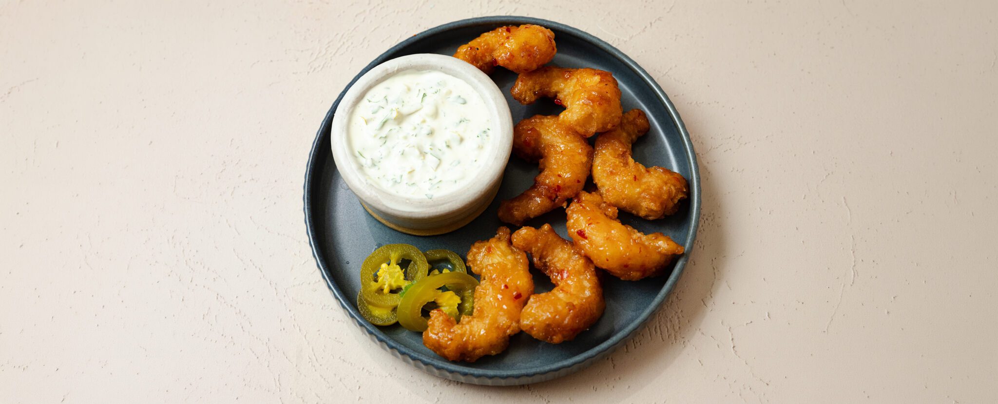 Three-quarter angle view of Social Kitchens Sweet & Spicy Shrimp on a round gray plate with a side of Jalapeño Cilantro Aioli in a white ramekin and a couple pieces of pickled jalapeño.