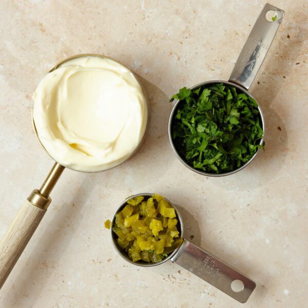 Overhead view of three metal measuring cups containing ½ cup mayo, ¼ cup pickled jalapeños, and ¼ cup chopped cilantro.