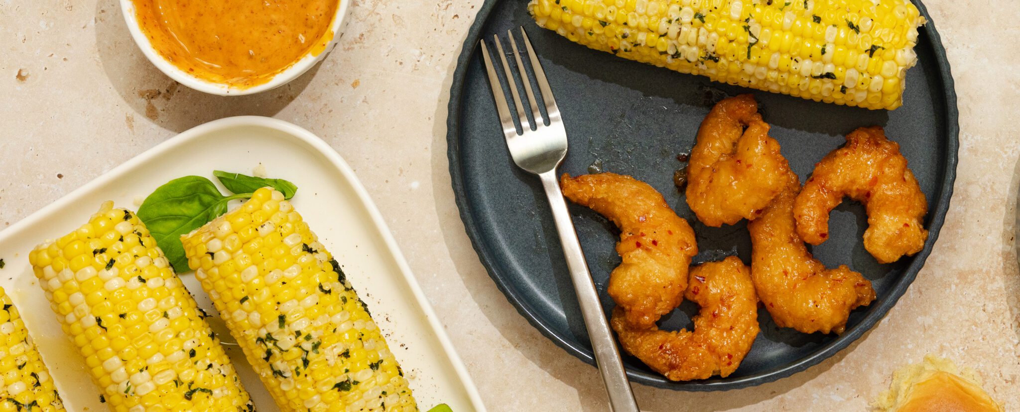 Overhead view of a gray plate with Social Kitchens Sweet & Spicy Shrimp with a cob of herby basil butter corn and a fork. Off to the side is a rectangular plate with several cobs of herby basil butter corn.
