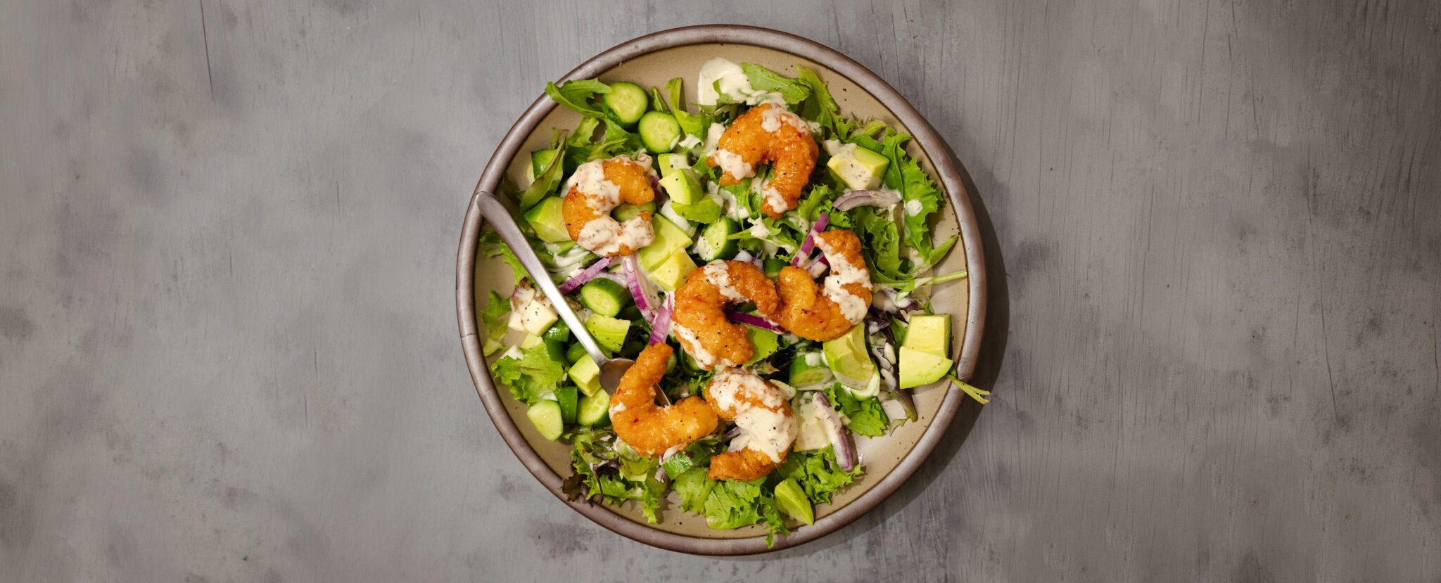 Overhead view of a No-Fuss Dinner Salad consisting of Social Kitchen Sweet & Spicy Shrimp, mixed greens, cucumber, diced avocado, red onion, and dressing.