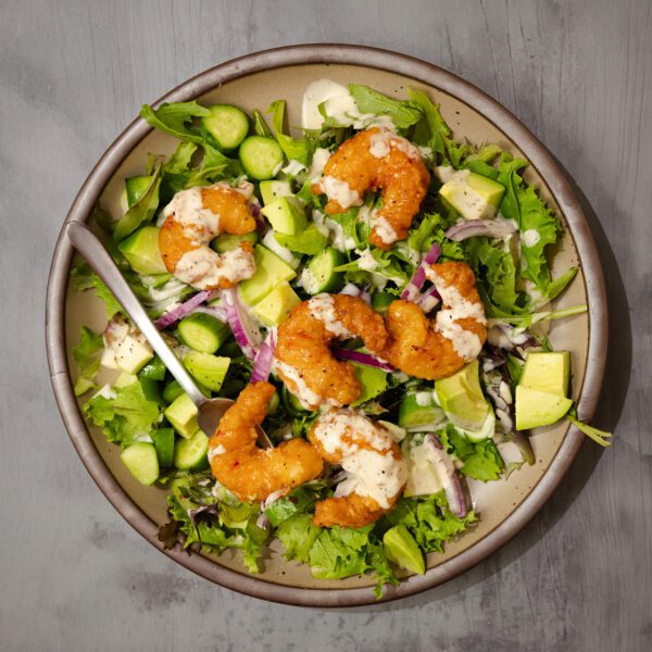 Overhead view of a No-Fuss Dinner Salad consisting of Social Kitchen Sweet & Spicy Shrimp, mixed greens, cucumber, diced avocado, red onion, and dressing.