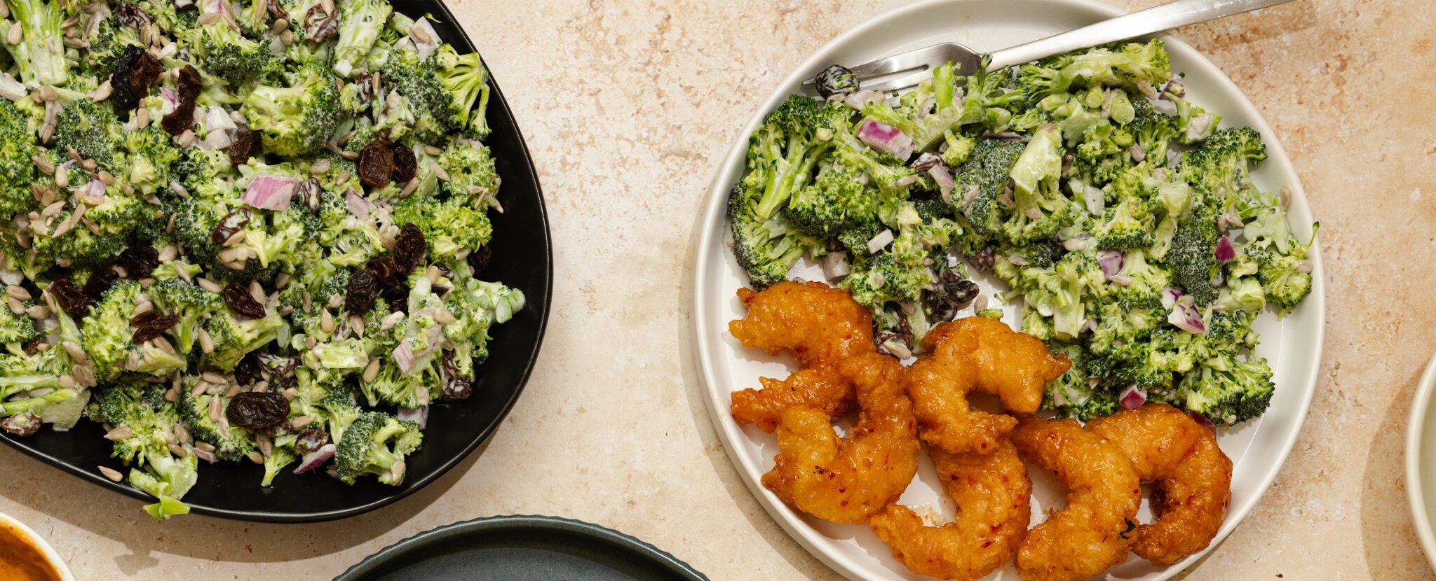 Overhead view of a black plate on the left with sweet & crunchy broccoli salad and a white plate on the right with the same salad and Social Kitchens Sweet & Spicy Shrimp.