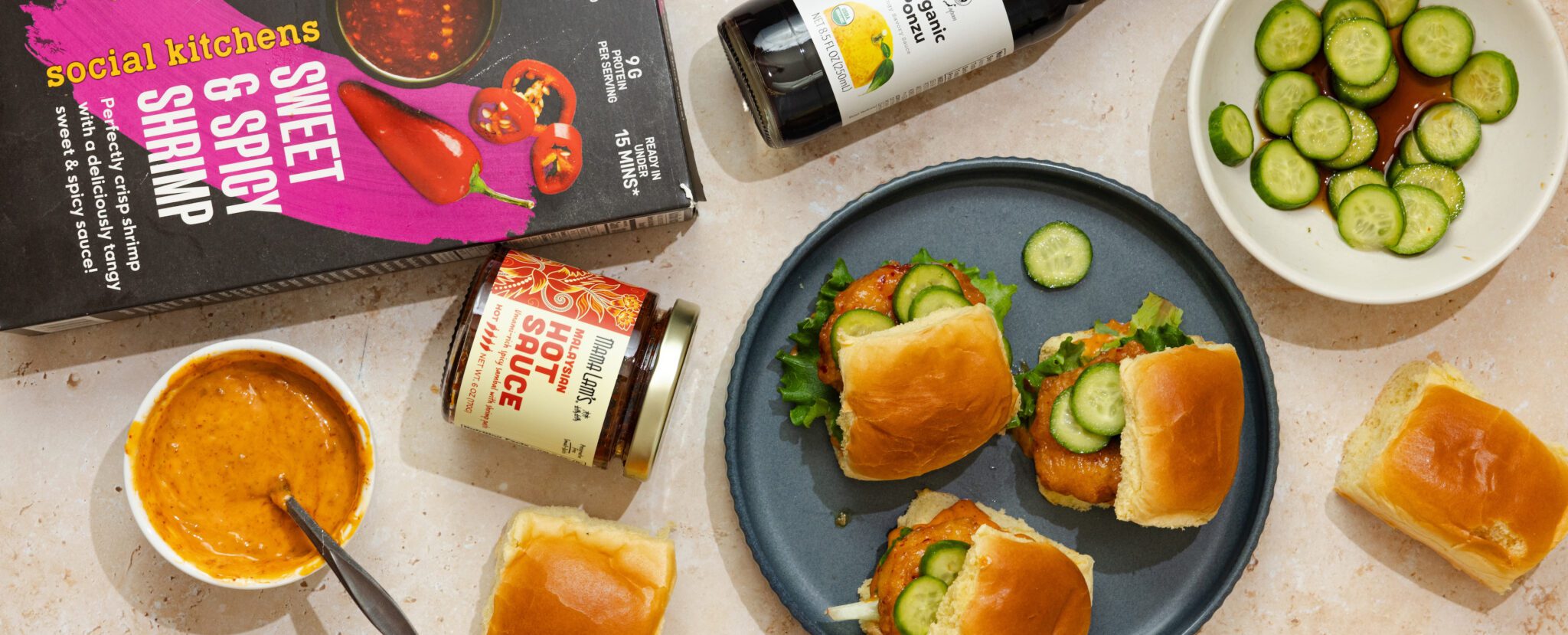 Overhead view of three Social Kitchens Sweet & Spicy Shrimp Sliders on a grey plate with a side of marinated sliced cucumbers, a bottle of Ponzu sauce, a jar of Mama Lam's Malaysian hot sauce, and Social Kitchens Sweet & Spicy Shrimp box packaging.