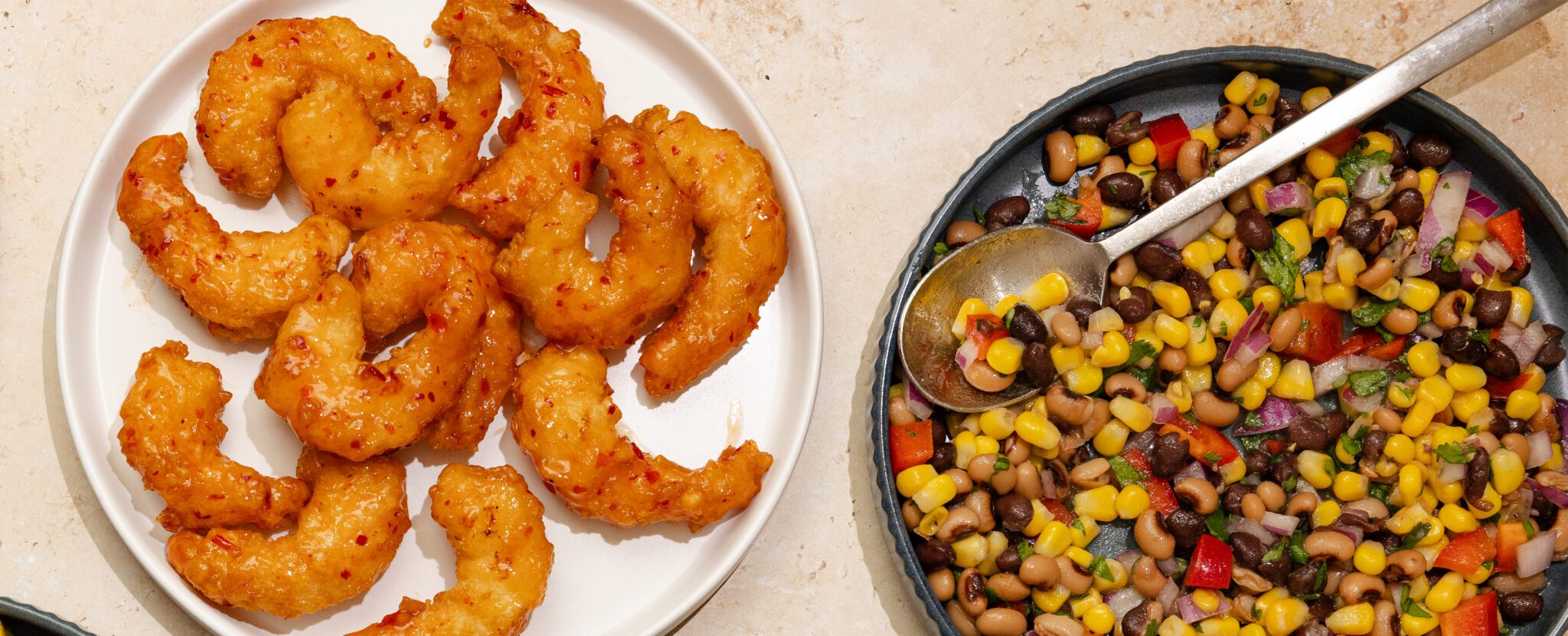 Overhead view of a black plate of Cowboy Caviar containing corn, black-eyed peas, red bell peppers, cilantro, and red onion. To the left is a white plate of Social Kitchens Sweet & Spicy Shrimp. Both plates are served over a beige stone table surface.
