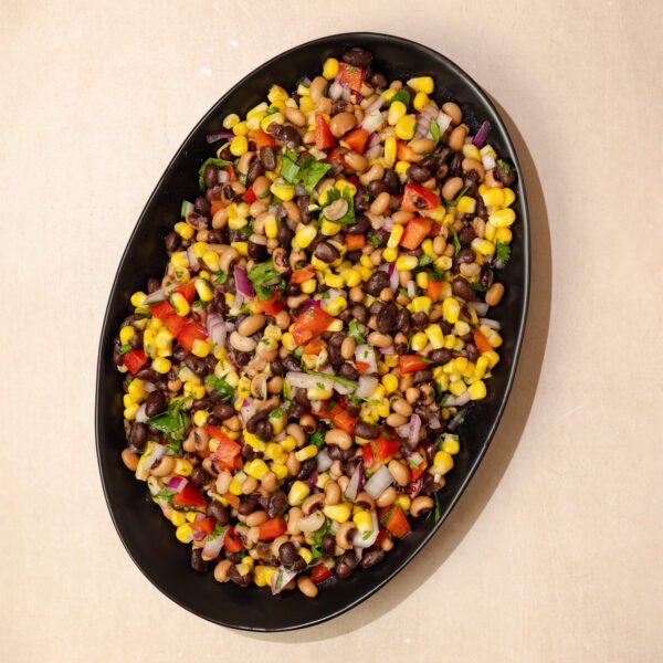 Overhead view of a large oval black plate of Cowboy Caviar which consists of corn, black-eyed peas, red bell peppers, cilantro, and red onion, served over a beige stone table surface.