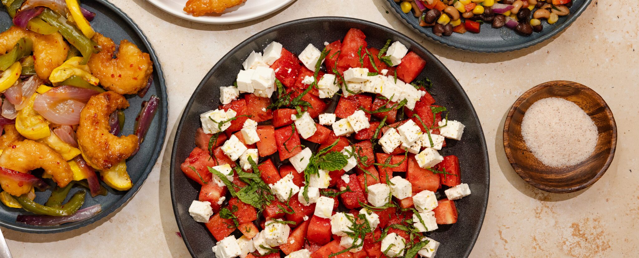 Overhead view of a Watermelon Feta Mint Salad on a round black plate, surrounded by an assortment of shrimp dishes and a wooden bowl of salt.