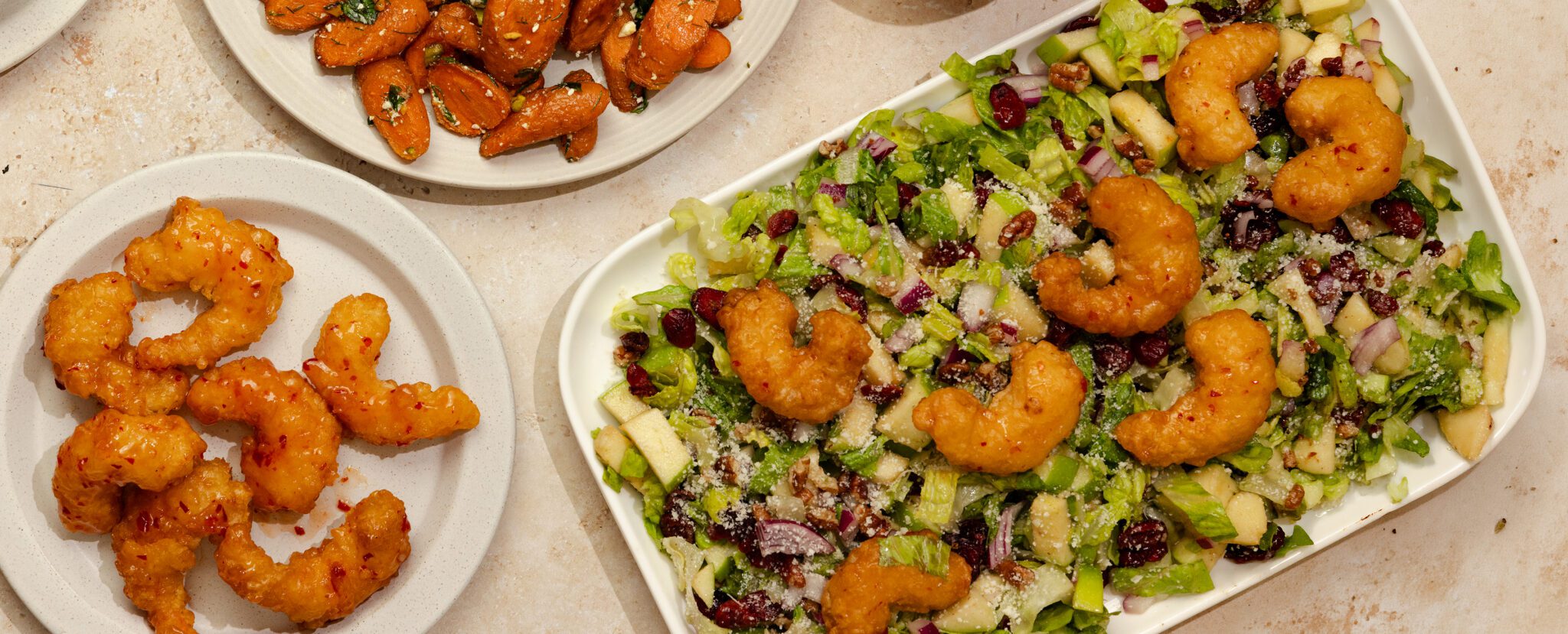 Overhead view of a large rectangular platter of Autumn Chopped Salad with Social Kitchens Sweet & Spicy Shrimp, a round plate of more shrimp, and another plate off to the side of roasted carrots.