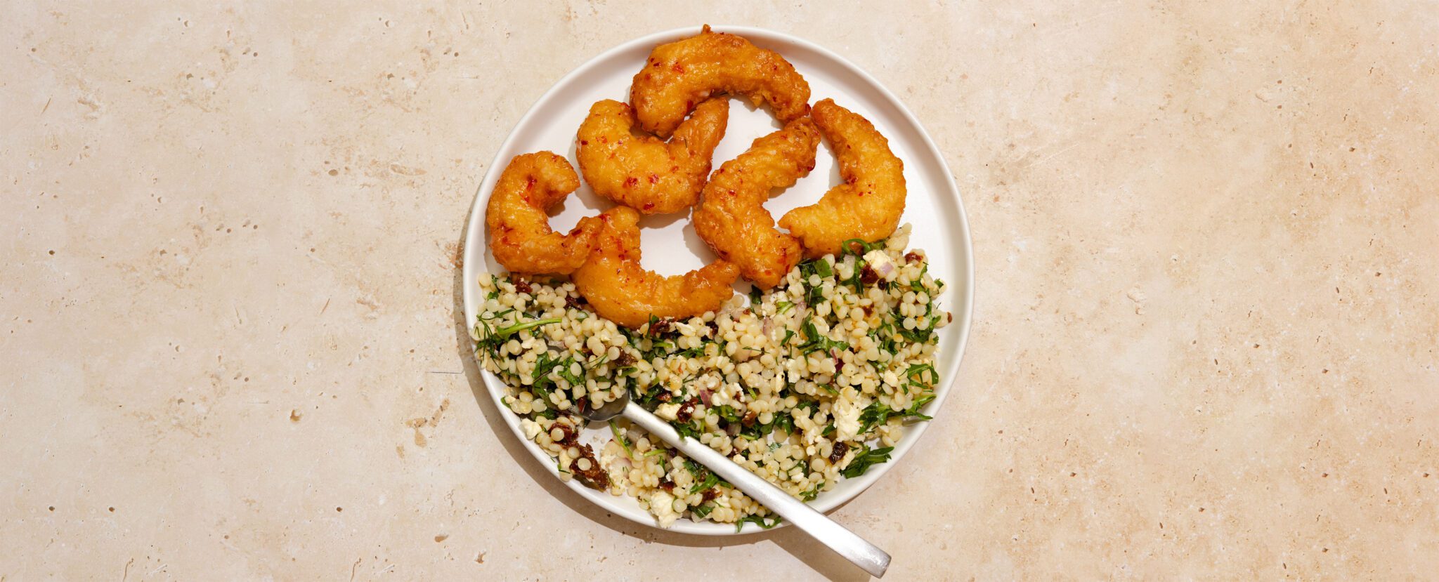 Overhead view of Social Kitchens Sweet & Spicy Shrimp with couscous pasta salad on a white plate with a fork, served over a beige stone table top.
