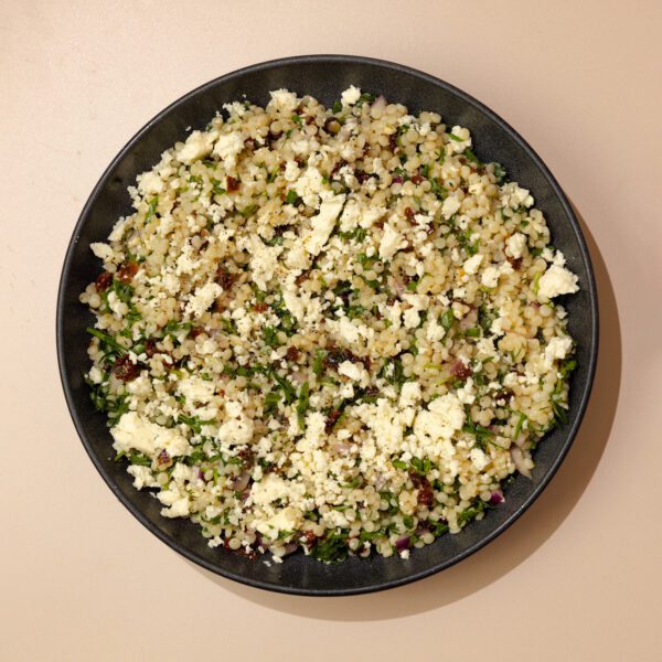 Overhead view of couscous pasta salad on a black plate over a beige stone table top.
