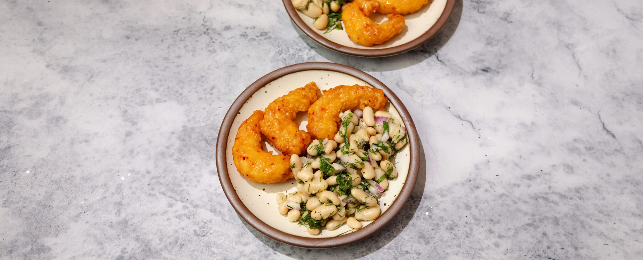 Three-quarter view of two plates each served with three pieces of Social Kitchens Sweet & Spicy Shrimp and a side of herby white bean salad over a marble table top.