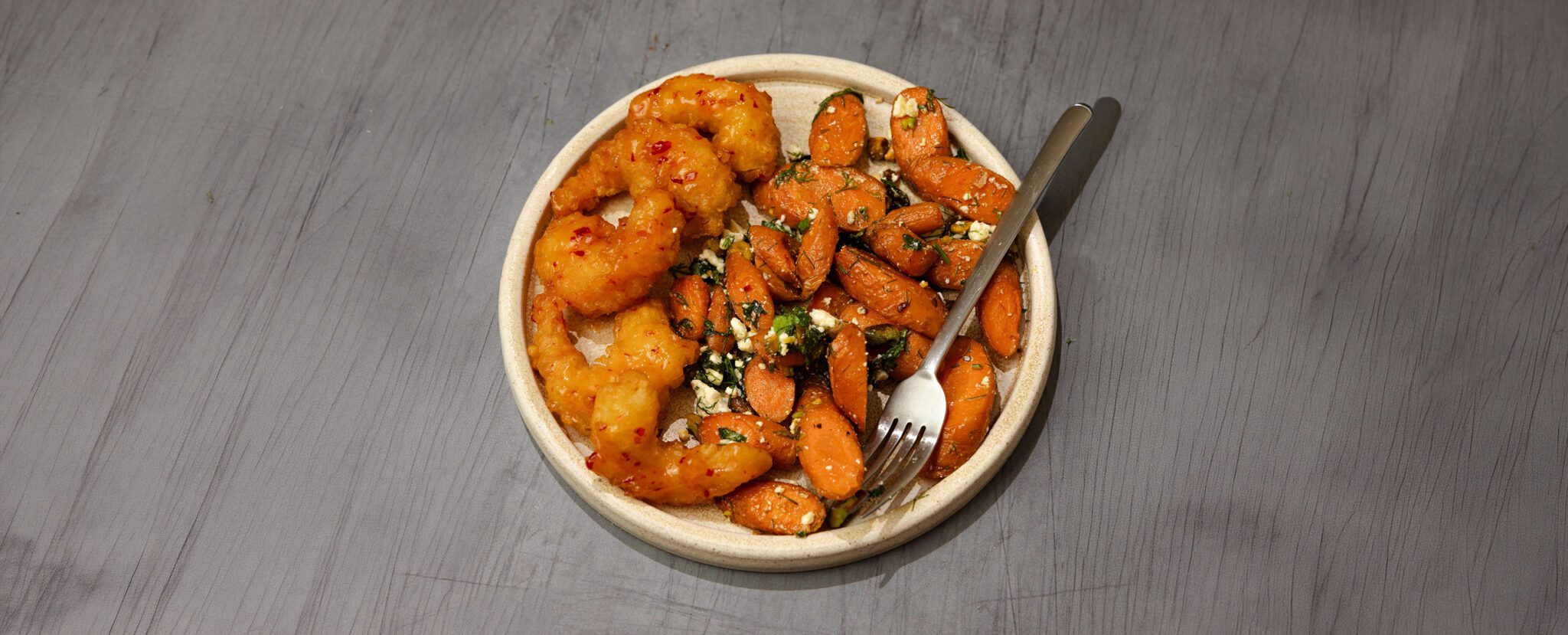 Three-quarter view of roasted herb feta & carrots with Social Kitchens Sweet & Spicy Shrimp on a round white plate with a fork, served over a gray wooden table top.