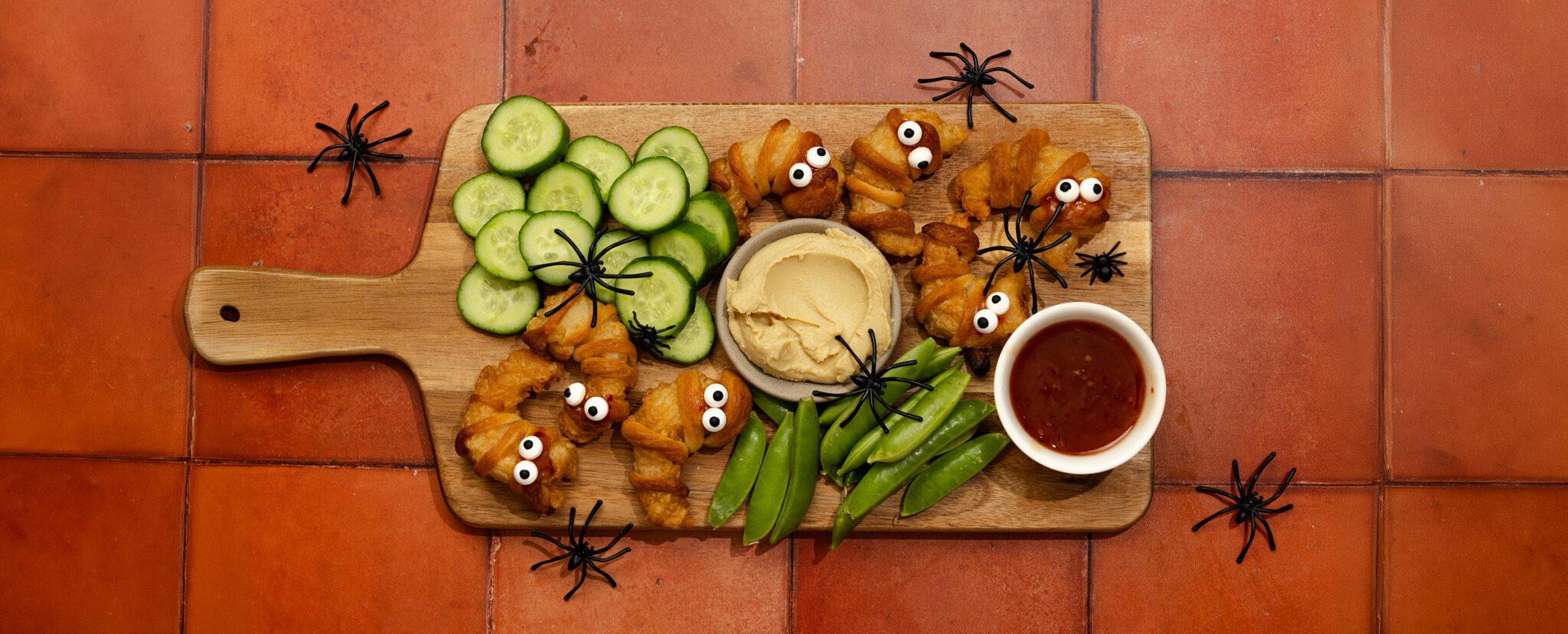 Overhead view of a charcuterie board spread of Spooky Shrimp Mummies made with Social Kitchens Sweet & Spicy Shrimp, sliced cucumbers, snap peas, hummus, and sweet & spicy sauce. Scattered throughout the spread of food and terracotta tiled tabletop are fake black spiders.