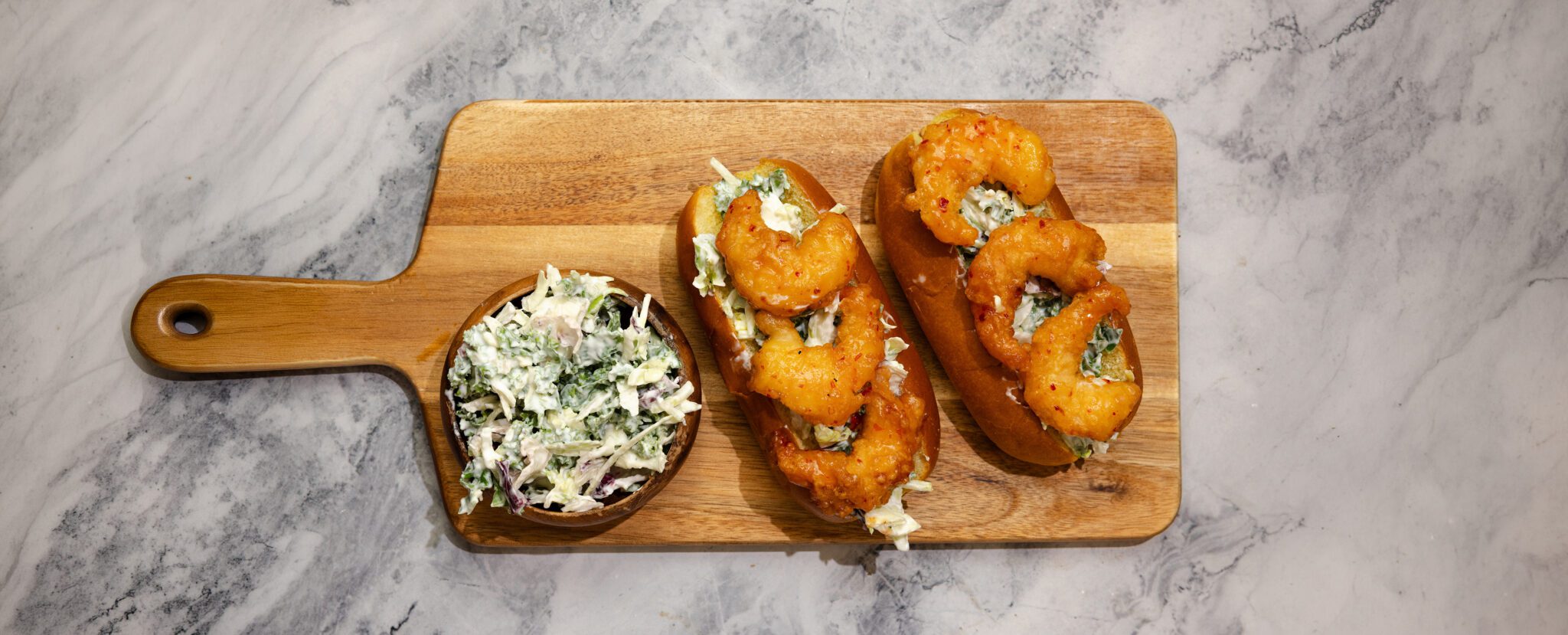Overhead view of Social Kitchen Sweet & Spicy Shrimp hoagie sandwiches served on a wooden cutting board with a side bowl of kale slaw over a marble table top.