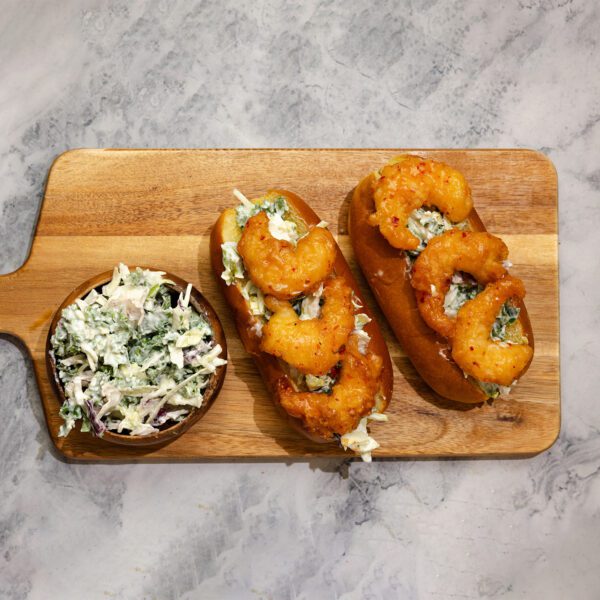 Overhead view of Social Kitchen Sweet & Spicy Shrimp hoagie sandwiches served on a wooden cutting board with a side bowl of kale slaw over a marble table top.